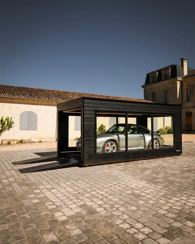 Garage Obsidian vu de côté dans la cour pavée d'un château, Porsche grise visible à travers les châssis vitrés